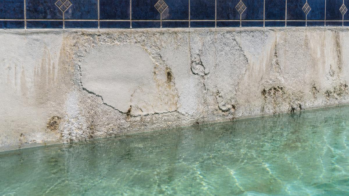 Close-up of a swimming pool interior wall showing visibly rough, stained, and flaking white plaster surface with waterline tile above, clear water and sunlight reflecting off the damaged finish